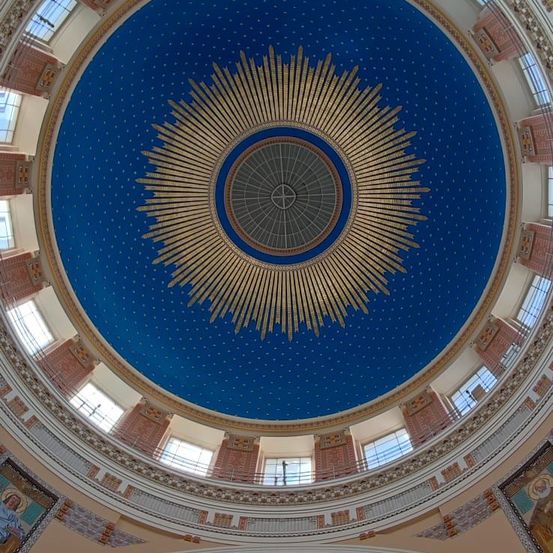 Eine runde blaue Decke mit goldenen Strahlen. In der Mitte der Strahlen befindet sich ein Kreuz. Die Decke hat ein blaues und goldenes Design. Rund um die Decke befinden sich Fenster.