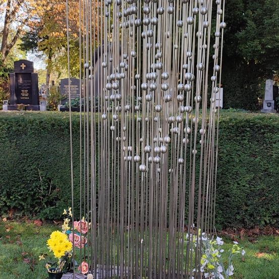 Eine metallische Skulptur mit zahlreichen silbernen Kugeln schmückt einen Friedhof. Hinter einer Hecke steht ein schwarzer Grabstein mit einem Kreuz. Vor der Skulptur sind frische Blumen platziert.