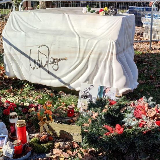 Ein bedecktes Grabmal mit einer Unterschrift, umgeben von Blumen, Kerzen und Kränzen auf einem Friedhof.