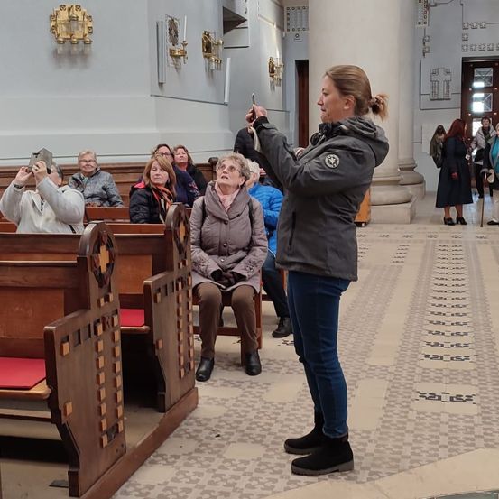 In einer Kirche steht eine Frau und macht ein Foto mit ihrem Handy, während mehrere Menschen auf Bänken sitzen. Einige schauen nach oben, andere beobachten sie. Der Innenraum hat weiße Säulen und einen gefliesten Boden.