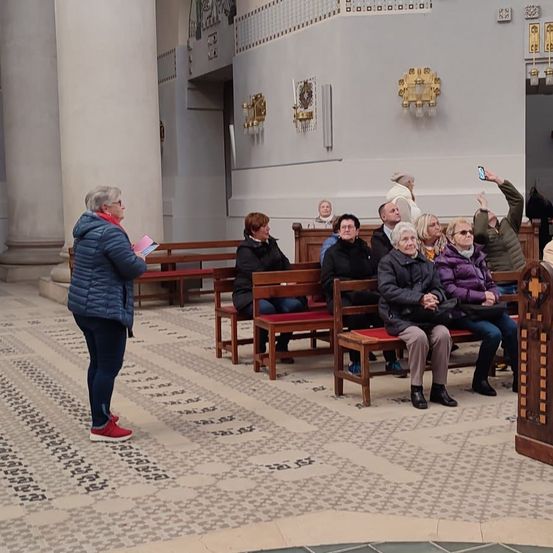 In einer Kirche sitzen eine Gruppe von Menschen auf Holzbänken. Eine Frau in einer blauen Jacke steht und hält ein Buch. Eine andere Frau macht ein Foto. Die Kirche verfügt über große Säulen und goldene Lampen an den Wänden.
