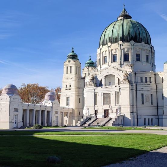 Ein großes weißes Gebäude mit einer grünen Kuppel, zwei Türmen und einer Treppe, die zum Eingang führt. Das Gebäude ist von grünem Gras und Bäumen umgeben.