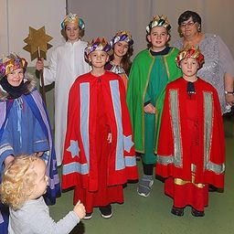 Eine Gruppe von Kindern und Erwachsenen in farbenfrohen Kostümen posiert für ein Foto in einem Raum. Sie halten Requisiten und lächeln.