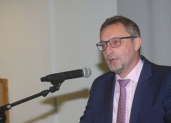 Ein Mann mit Brille und Anzug steht an einem Rednerpult und spricht in ein Mikrofon. Die Wand hinter ihm ist weiß, und links befindet sich eine Holztür.