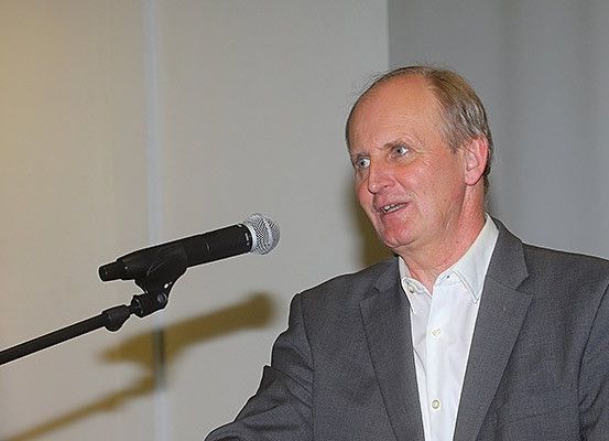 Ein Mann steht an einem Rednerpult mit einem Mikrofon, gekleidet in einem grauen Anzug und einem weißen Hemd, spricht in einer Innenumgebung.