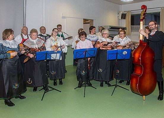 Eine Gruppe Musiker, gekleidet in traditioneller Kleidung, steht in einem Raum und spielt verschiedene Instrumente. Ein Mann steht rechts und hält einen großen Kontrabass.