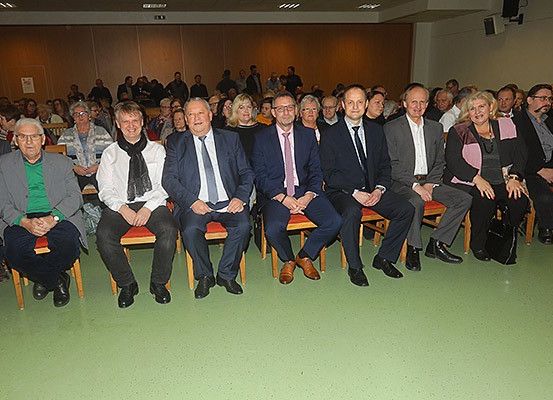 Eine Gruppe von Menschen sitzt in Reihen in einem Raum, einige in formeller Kleidung. Sie nehmen wahrscheinlich an einer Versammlung teil. Der Raum hat Holzwände und einen grünen Boden.
