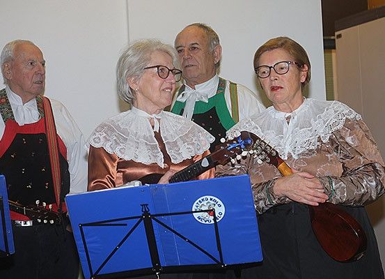 Vier ältere Personen, zwei Männer und zwei Frauen, stehen vor einer weißen Wand. Sie tragen alle eine Brille und traditionelle Kleidung. Die Frau auf der rechten Seite hält ein Musikinstrument. Der Mann auf der linken Seite hält eine Gitarre. Die Frau in der Mitte hält eine Gitarre und ein Musikinstrument.