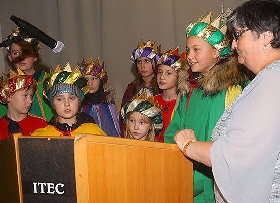 Kinder in festlichen Kostümen stehen um ein Podium herum, eines trägt eine Krone, wahrscheinlich an einer Aufführung beteiligt. Eine Frau steht in der Nähe und beobachtet sie.
