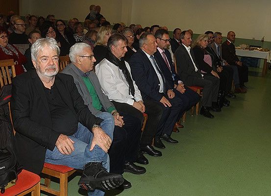 Eine Gruppe von Menschen in formeller Kleidung sitzt in einer Reihe von Stühlen in einer geschlossenen Umgebung, wahrscheinlich bei einer Veranstaltung. Ein Mann in einem schwarzen Mantel und Jeans sitzt in der vorderen Reihe, während eine schwarze Kameratasche auf dem Stuhl neben ihm steht.