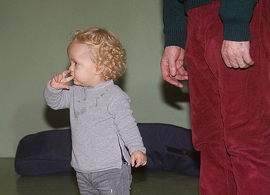 Ein kleiner Junge mit blonden Locken steht, Hand am Mund, und sieht einen Erwachsenen an, der eine schwarze Strickjacke und rote Hosen trägt.