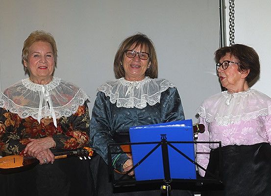 Drei Frauen in traditioneller Kleidung, lächelnd und mit Saiteninstrumenten, stehen vor einer weißen Wand mit einem Notenständer zwischen ihnen.