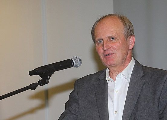 Ein Mann in einem grauen Anzug und weißem Hemd steht an einem Mikrofon und spricht wahrscheinlich bei einer Veranstaltung oder Konferenz. Er hat einen freundlichen Ausdruck.