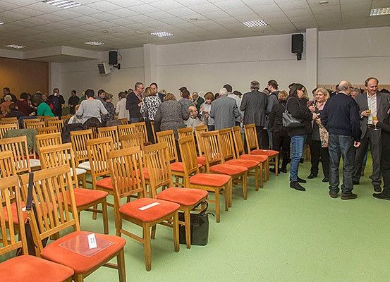 Menschen stehen in Reihen von Stühlen mit roten Sitzen in einem Raum. Sie halten Getränke in den Händen. Einige unterhalten sich. Der Raum hat eine Holzwand, eine weiße Decke und einen grünen Boden.