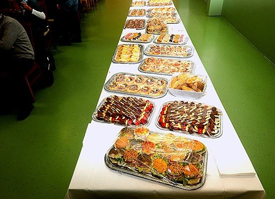 Ein langer Tisch ist mit einer weißen Tischdecke bedeckt. Es gibt Schalen mit verschiedenen Desserts, darunter Gebäck, Kuchen und Sandwiches. Der Boden ist grün, und eine Person sitzt auf einem Stuhl im Hintergrund.