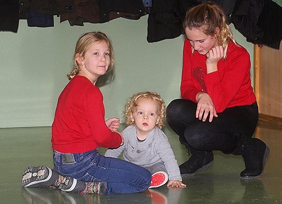 Zwei junge Mädchen und ein Kleinkind sitzen auf dem Boden. Ein Mädchen hält die Hand des Kleinkindes, während ein anderes kniet und mit ihm spricht. Sie befinden sich in einem Raum mit einer grünen Wand und Kleidern, die an einem Kleiderständer hängen.