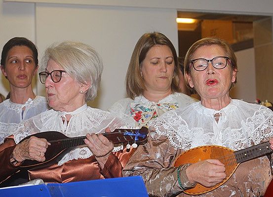 Eine Gruppe älterer Frauen in traditioneller Kleidung spielt Saiteninstrumente, möglicherweise Mandolinen. Sie stehen eng beieinander, eine Frau singt, während eine andere nach unten schaut. Die Frau auf der rechten Seite trägt eine Brille. Sie scheinen an einer musikalischen Veranstaltung teilzunehmen.