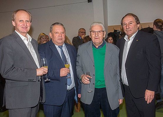 Vier Männer in Anzugkleidung stehen lächelnd nebeneinander, mit Weinläsern in den Händen, während eine Menschenmenge im Hintergrund bei einer Veranstaltung steht.