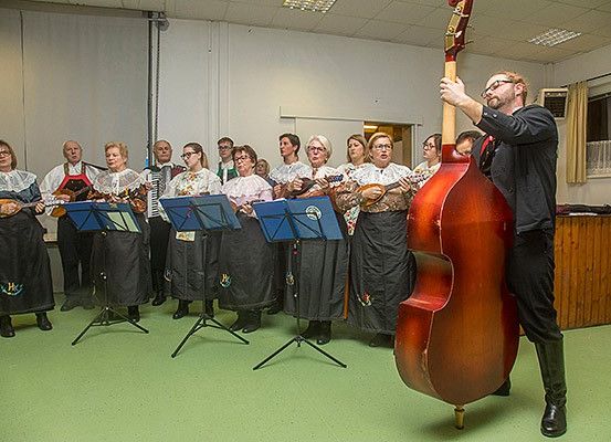 Eine Gruppe von Musikern in traditioneller Kleidung spielt verschiedene Instrumente in einem Raum mit grünem Boden. Ein Mann steht mit einem Cello.
