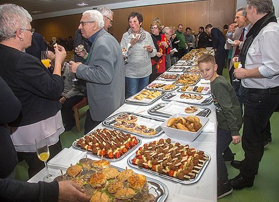 Eine Gruppe von Menschen versammelt sich um einen langen Tisch mit verschiedenen Speisen, darunter Gebäck, Sandwiches und Spieße. Sie scheinen das Buffet zu genießen.