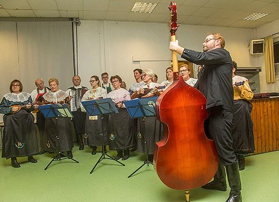 Eine Gruppe von Musikern tritt in einem Raum mit grünem Boden auf. Sie halten verschiedene Instrumente, darunter ein Cello, und einige spielen Akkordeon. Sie sind in traditioneller Kleidung gekleidet.