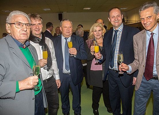 Eine Gruppe von Männern und Frauen in formeller Kleidung, die Weinglas halten und lächeln, möglicherweise bei einer gesellschaftlichen Veranstaltung.