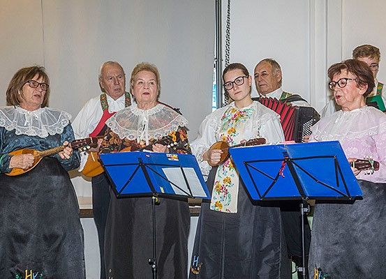 Eine Gruppe älterer Musiker in traditionellen Kostümen steht vor Musikständen und spielt Saiteninstrumente. Sie befinden sich in einem Raum mit einer weißen Wand im Hintergrund.