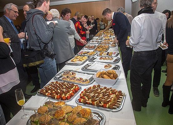 Eine Versammlung von Menschen an einem Buffet mit verschiedenen Speisen auf Tabletts, einige stehen und andere unterhalten sich, halten Getränke und Teller in der Hand, drinnen.