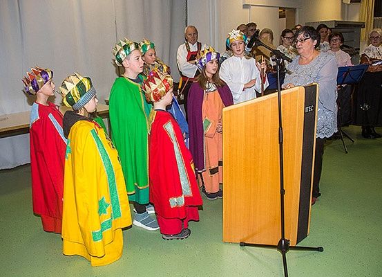 Kinder in bunten Kostümen stehen vor einem Podium, wahrscheinlich bei einer Schulveranstaltung, während eine erwachsene Frau in ein Mikrofon spricht.