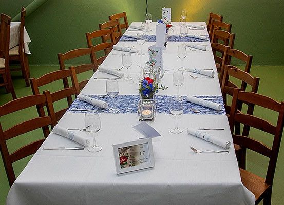Ein langer Esstisch mit blauen Tischläufern ist mit weißen Tischdecken, Weingläsern und Besteck gedeckt. Auf dem Tisch befinden sich eine Kerze und ein Bilderrahmen. Der Raum hat grüne Wände.