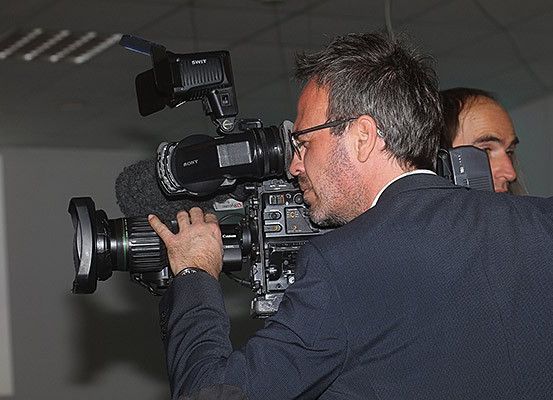 Ein Mann in einem Anzug hält eine Sony-Videokamera und scheint zu filmen. Eine weitere Person steht hinter ihm.