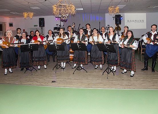 Eine Gruppe Musiker, gekleidet in traditioneller Kleidung, steht auf einer Bühne mit Saiteninstrumenten und singt und spielt Musik.