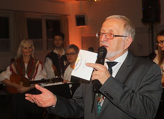 Ein älterer Mann hält ein Mikrofon in der Hand und spricht vor einer Gruppe von Menschen. Er trägt eine Brille und einen Anzug. Hinter ihm spielt eine Frau Gitarre.