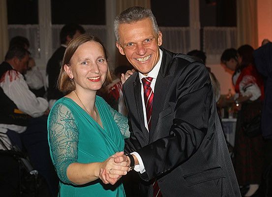 Ein Mann und eine Frau tanzen bei einer Veranstaltung, lächeln und halten sich an den Händen. Die Frau trägt ein grünes Kleid und der Mann einen schwarzen Anzug. Hinter ihnen sind andere Personen anwesend, einige sitzend und andere stehend.