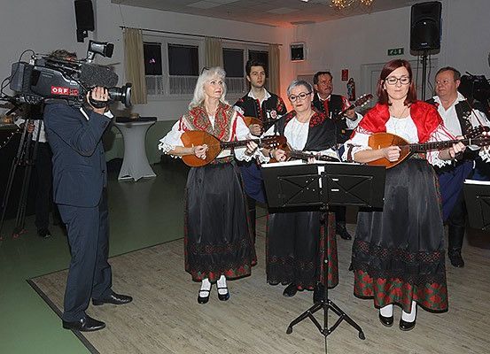Eine Gruppe von Menschen in traditioneller Kleidung spielt Saiteninstrumente auf der Bühne. Ein Kameramann filmt sie.