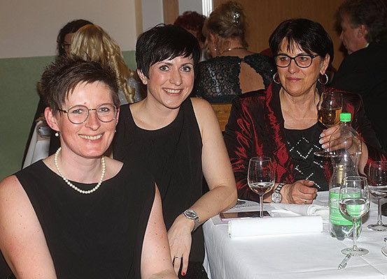 Drei Frauen sitzen bei einem formellen Event lächelnd an einem Tisch. Sie tragen schwarze Kleider und Brillen. Die Frau auf der rechten Seite hält eine Flasche Wein und ein Weinglas. Hinter ihnen sitzen weitere Personen, die in Gespräche vertieft sind.