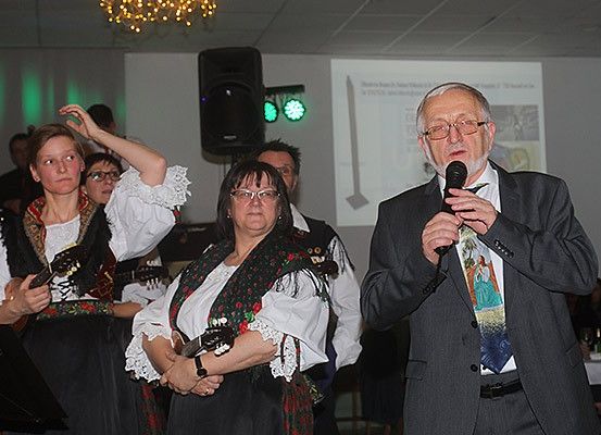 Ein älterer Mann spricht in ein Mikrofon, während eine Frau in traditioneller Kleidung Gitarre spielt.