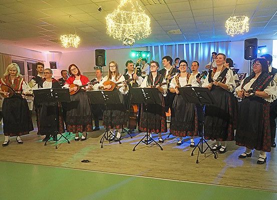 Eine Gruppe von Menschen in traditioneller Kleidung steht auf der Bühne und spielt Saiteninstrumente in einem Raum mit Kronleuchtern und Lautsprechern.