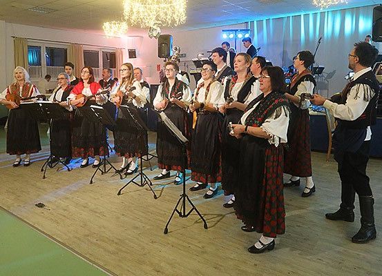 Eine Gruppe von Menschen in traditioneller Kleidung steht auf der Bühne, hält Gitarren und singt. Sie werden von einer Band begleitet, und die Szene spielt sich drinnen ab.