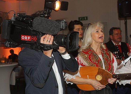 Ein Mann filmt eine Frau, die mit einem traditionellen Instrument singt, während andere zusehen. Dahinter steht ein Tisch mit Gläsern und einem Lautsprecher.