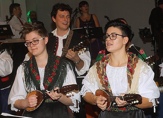 Eine Gruppe von Musikern in traditioneller Kleidung spielt verschiedene Saiteninstrumente, während einige Leute dahinter stehen und zusehen. Die Musiker halten Gitarren und Mandolinen.
