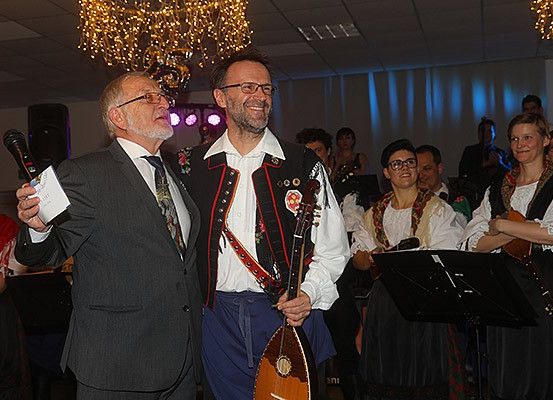 Zwei Männer stehen vor einem Mikrofon und einer Gruppe von Menschen. Der Mann auf der linken Seite hält ein Mikrofon und Papier und lächelt. Der Mann neben ihm hält ein Laouto. Hinter ihnen spielt eine Frau Geige. Eine Krone leuchtet über ihnen.