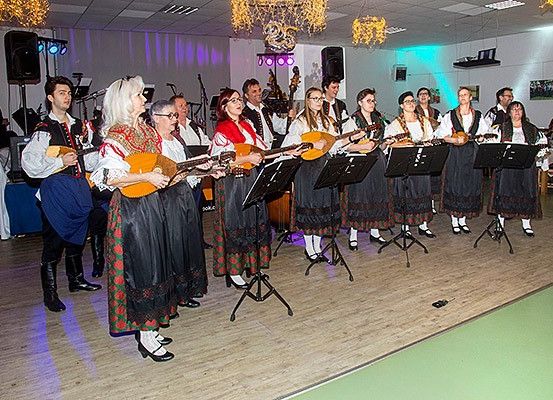 Eine Gruppe von Musikern in traditioneller Kleidung tritt auf einer Bühne auf und spielt Lauten. Sie stehen vor Notenständern. Die Bühne ist gut beleuchtet, und Kronleuchter hängen von der Decke.
