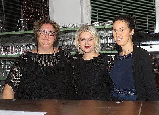 Drei Frauen stehen gemeinsam hinter einer Bar und lächeln für ein Foto. Die Frau auf der linken Seite trägt eine Brille und eine Halskette. Die Frau in der Mitte hat blonde Haare und eine Blume auf ihrem Kleid. Die Frau auf der rechten Seite trägt ein blaues Oberteil und eine schwarze Strickjacke. Hinter ihnen befindet sich ein Regal mit Gläsern und ein Fenster mit Jalousien.