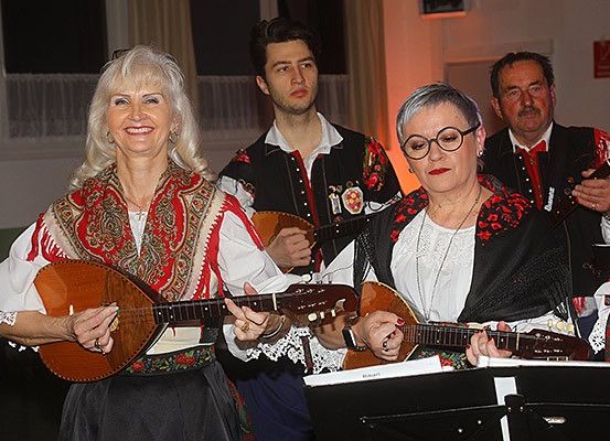 Fünf Menschen in traditionellen Kostümen spielen Saiteninstrumente in einem Raum. Die Frau links lächelt in die Kamera. Die andere Frau trägt eine Brille. Hinter ihnen schaut ein Mann in die Kamera. Vor ihnen steht ein Notenständer.