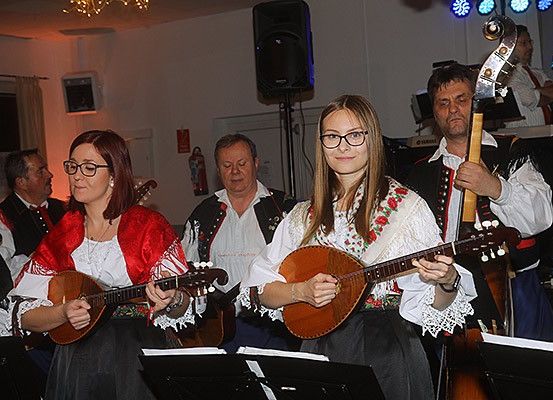 Eine Gruppe von Menschen in traditionellen Kostümen spielt in einem Raum Musikinstrumente. Die beiden Frauen spielen Lauten. Ein Mann spielt Cello. Hinter ihnen steht ein Lautsprecher und ein Fenster mit einem Vorhang. Hinter ihnen stehen weitere Menschen, die wahrscheinlich auch Instrumente spielen.