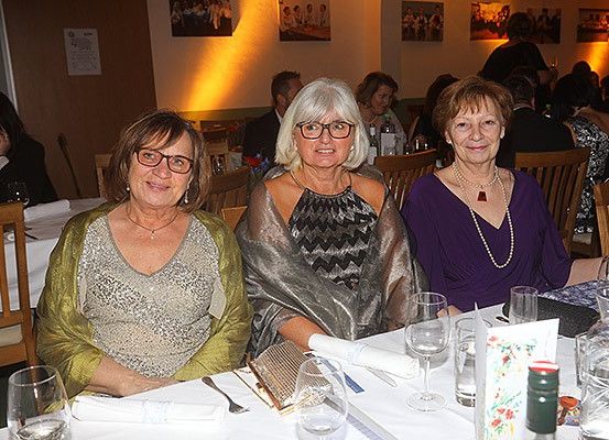 Drei ältere Frauen sitzen an einem Tisch und lächeln für ein Foto. Sie tragen Brillen und Schmuck, mit Weingläsern und einer Flasche in der Nähe. Andere Leute sitzen an Tischen hinter ihnen, und Bilder hängen an der Wand.