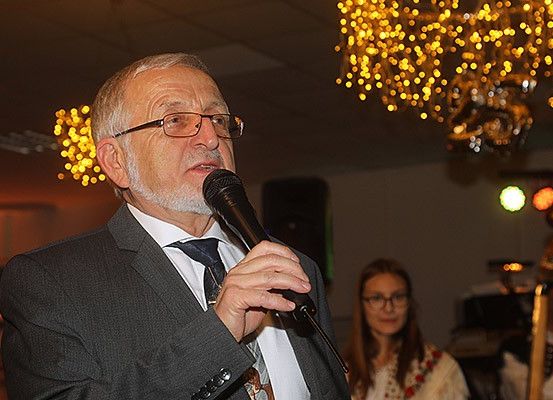 Ein älterer Mann in einem Anzug spricht in ein Mikrofon, während er vor einer Frau und einer Kristallleuchte steht.