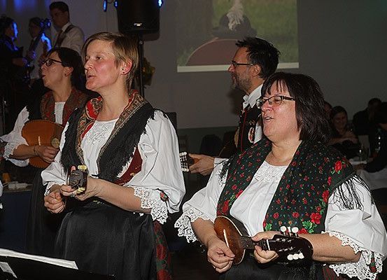 Mehrere Menschen treten in einem Raum auf. Sie spielen traditionelle Saiteninstrumente. Die Frau rechts hält eine Mandoline. Ein Beamer ist an der Wand montiert.