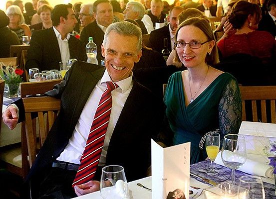 Ein Mann und eine Frau sitzen zusammen an einem Tisch mit Getränken und Blumen und lächeln für ein Foto in einem formellen Ambiente.